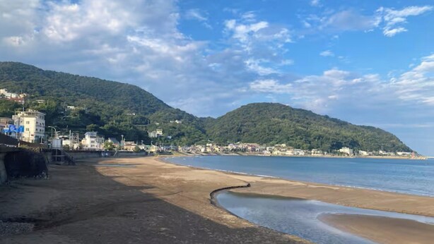 Una playa con una costa curva, edificios en una colina y un cielo despejado.