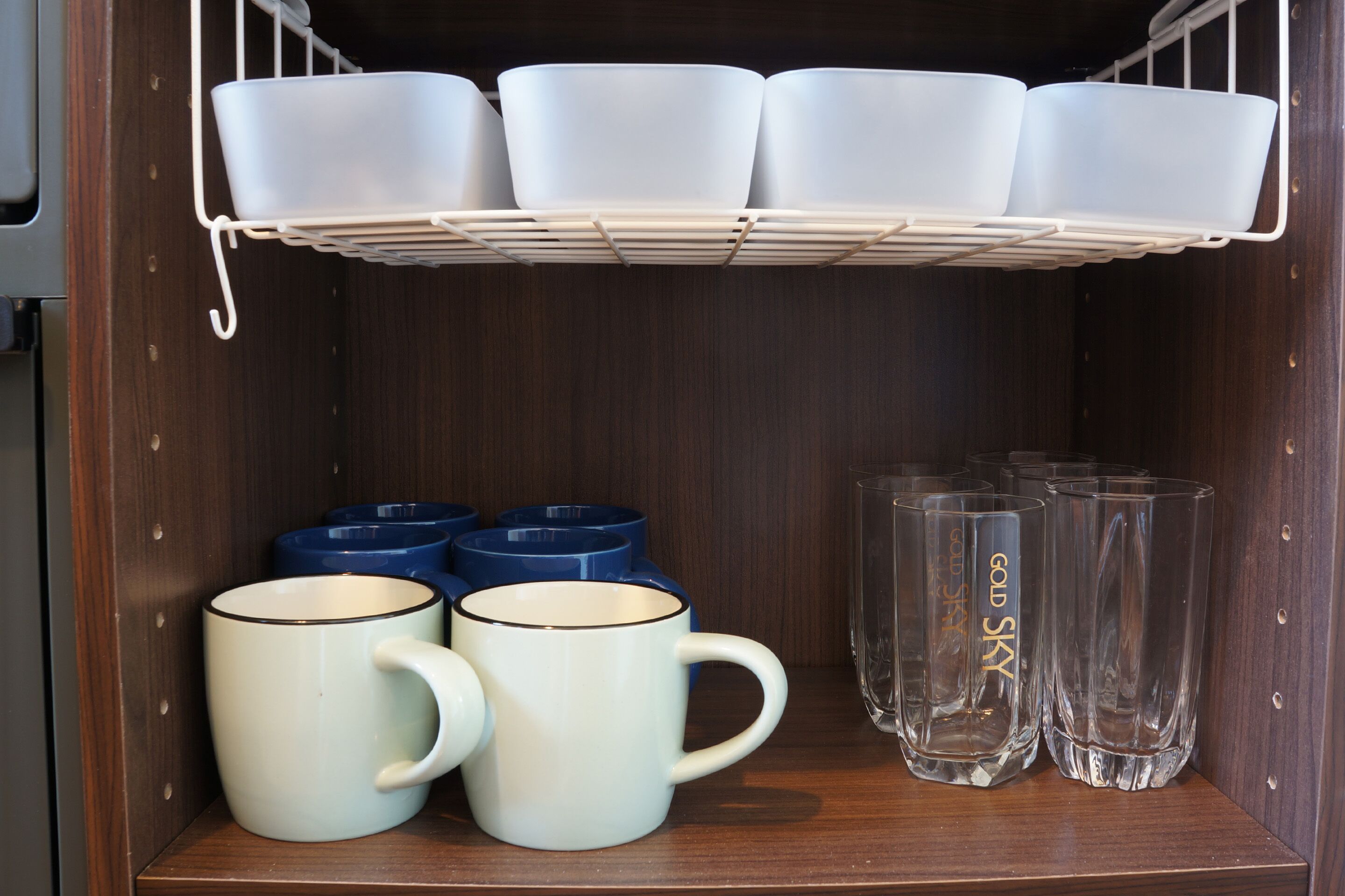 Una repisa con tazas blancas y azules, una bandeja blanca con tazas blancas, y vasos transparentes con texto dorado.