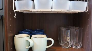 Una repisa con tazas blancas y azules, una bandeja blanca con tazas blancas, y vasos transparentes con texto dorado.