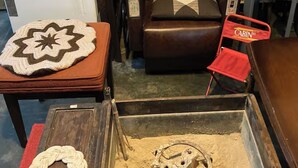 Una habitación con un sofá de cuero marrón, una silla plegable roja, una mesa de madera con un mantel tejido y una bandeja metálica con un objeto decorativo.