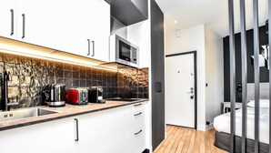 A modern kitchen with white cabinets, a microwave, and a coffee maker. There is a wooden floor and a door leading to a bedroom with a bed.