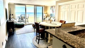 A spacious room with a large window overlooking the ocean, a dining area with a round table, and a kitchen with granite countertops.