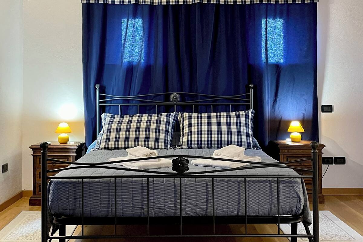 Un dormitorio con estructura de cama metálica, colcha a cuadros, dos lámparas de noche y una ventana con cortinas azules.