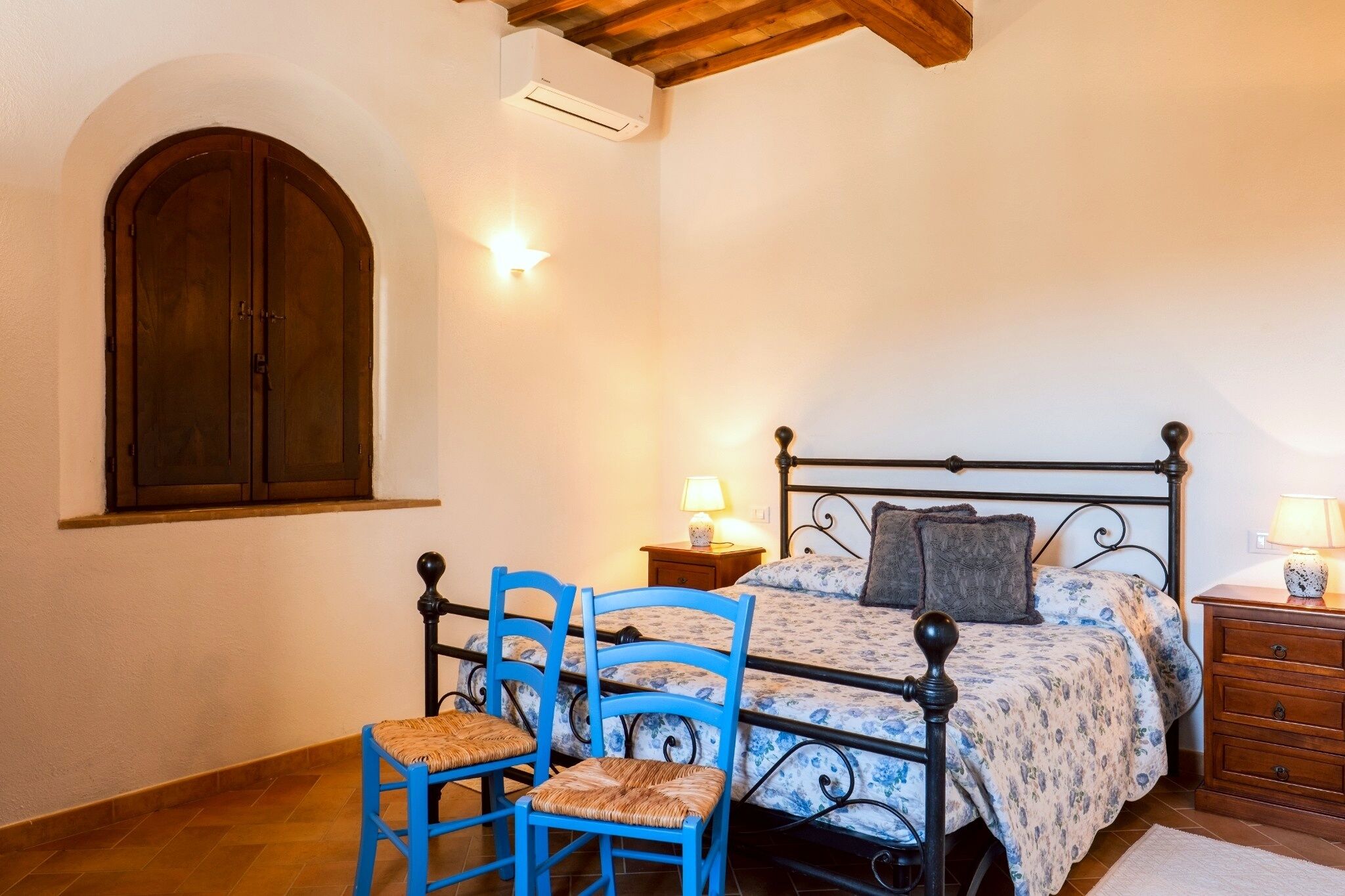 A bedroom with a bed, two nightstands, a chair, a wooden cabinet, and a window with an arched design.