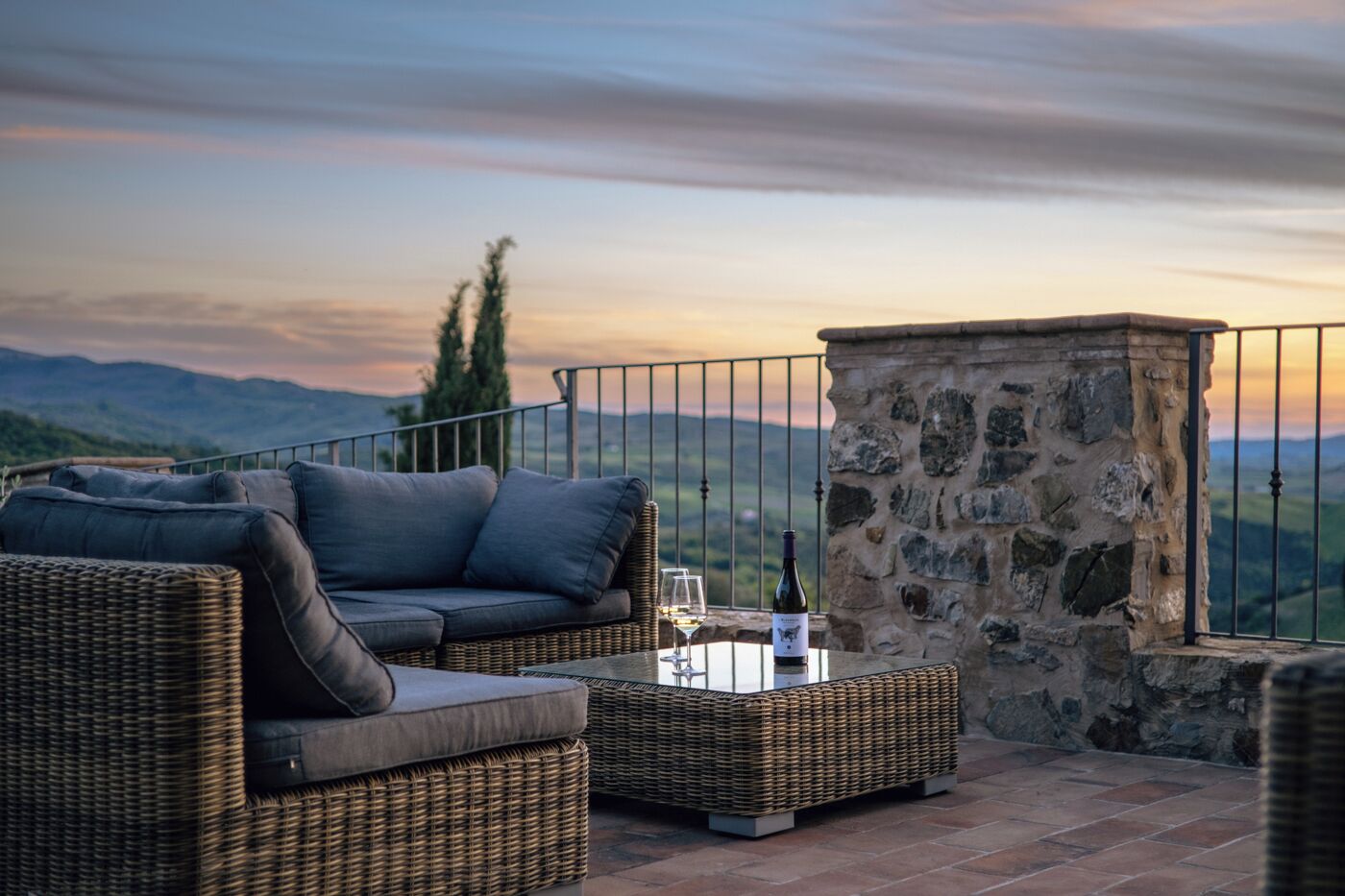 A wicker sofa and table set on a terrace with a stone wall and a view of the hills.