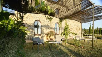 A stone building with arched windows, a covered outdoor seating area with chairs, and a pergola with climbing plants.