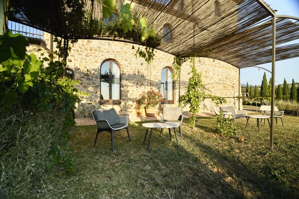 Un edificio de piedra con ventanas arqueadas, un espacio al aire libre cubierto con sillas, y una pérgola con plantas trepadoras.