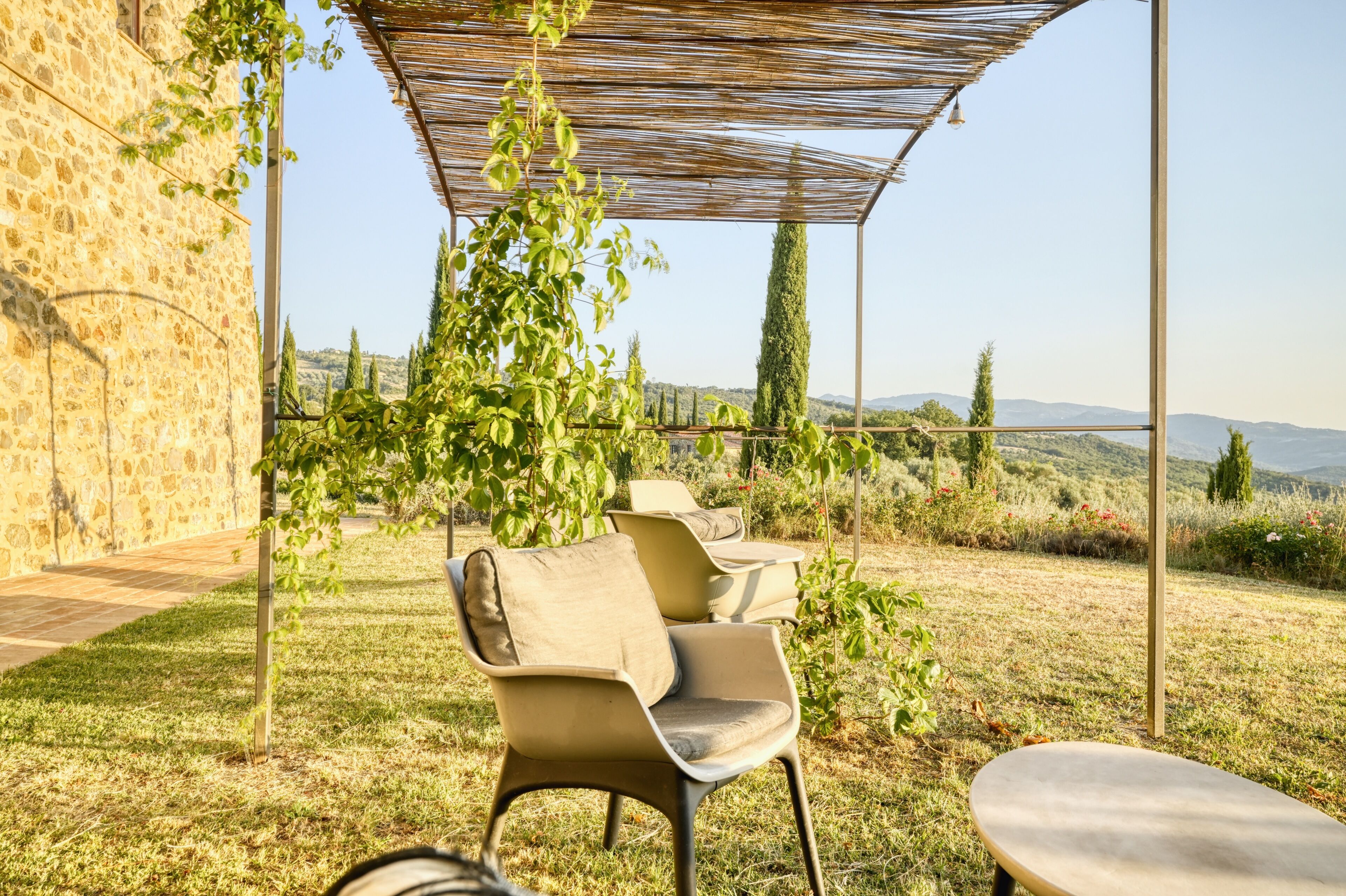 Un espacio al aire libre con sillas, una mesa, una pérgola y una pared de piedra.