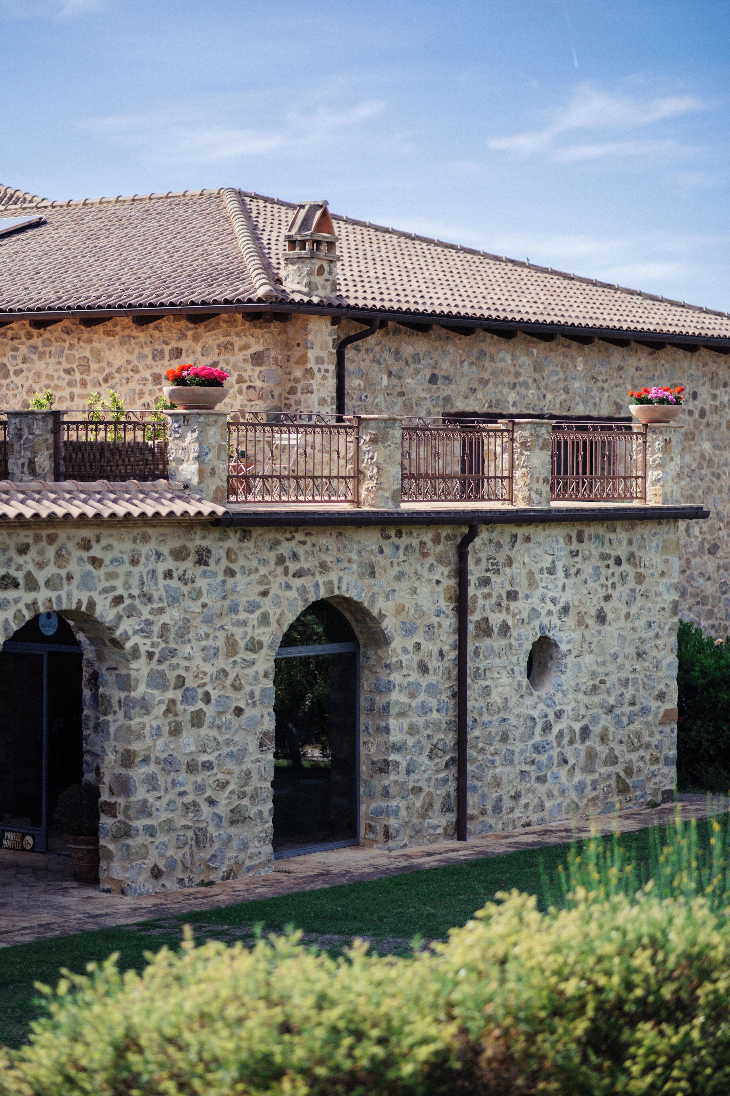 A stone building with a tiled roof, arched windows, and a balcony with flower pots.