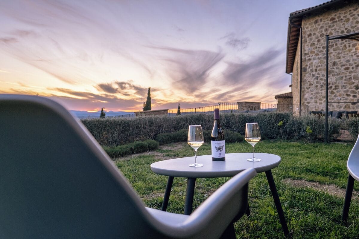 A table set outdoors with two wine glasses and a bottle, surrounded by greenery and a stone building in the background.