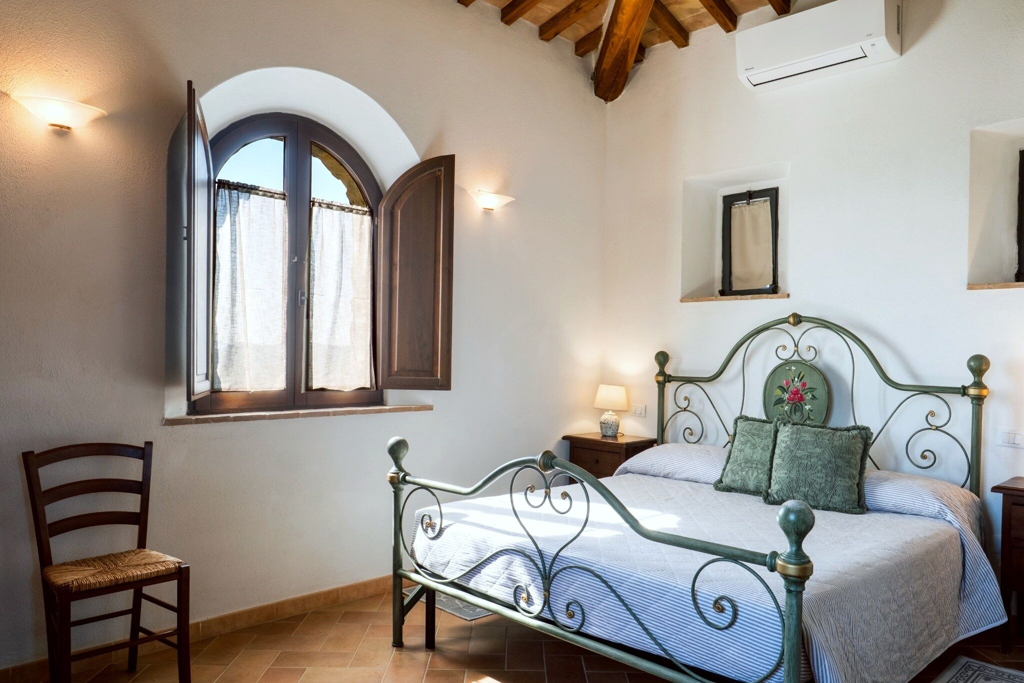 Une chambre à coucher comprenant un lit, une chaise, une table de chevet, une fenêtre avec des rideaux et un tableau accroché au mur.