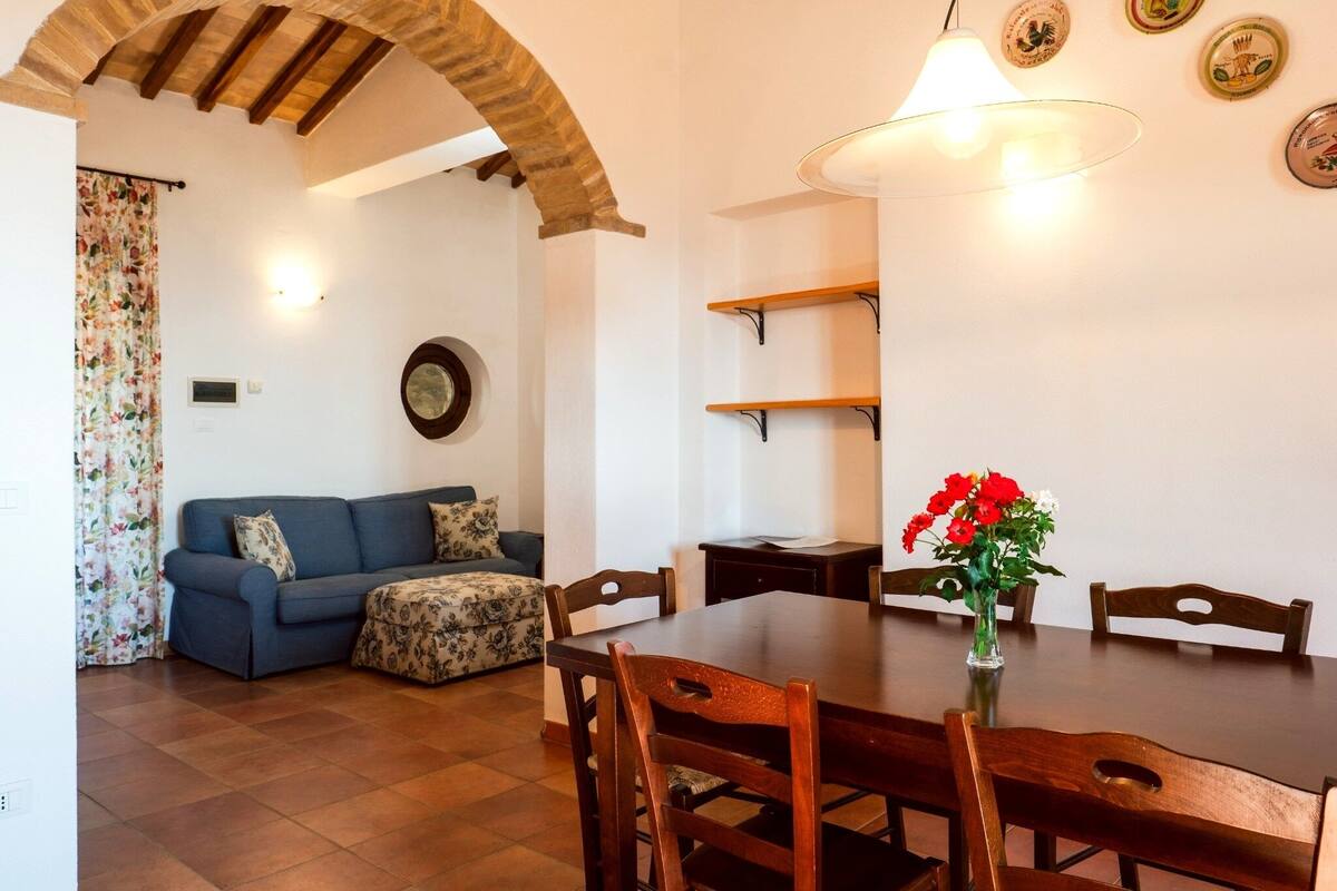 Un comedor con una mesa y sillas de madera, un jarrón con flores rojas y un sofá azul con cojines estampados.