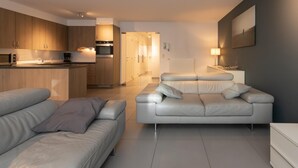 A modern living room with a sofa and a white leather couch, a kitchen with wooden cabinets and a built-in oven, and a hallway leading to a dining area. - Apartment in Ostend near the Seafront (OSTEND)