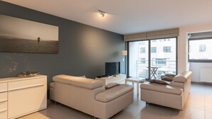 A modern living room with a large window, a flat-screen TV, and a white sofa. - Apartment in Ostend near the Seafront (OSTEND)