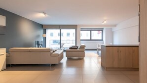 A modern living room with a sofa, a smaller seating area, and a kitchen area with wooden cabinets. - Apartment in Ostend near the Seafront (OSTEND)