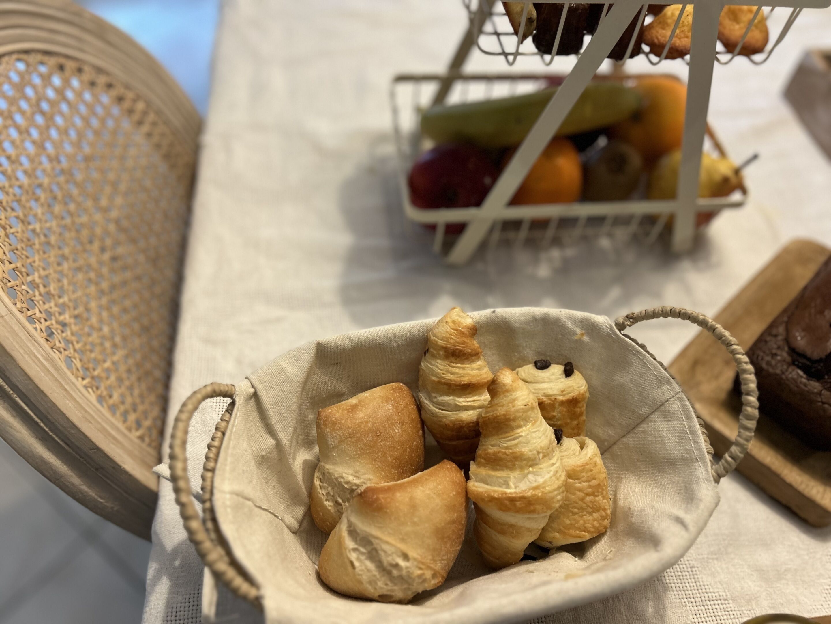 Een mandje croissants op een tafel, met een fruitmand op de achtergrond.