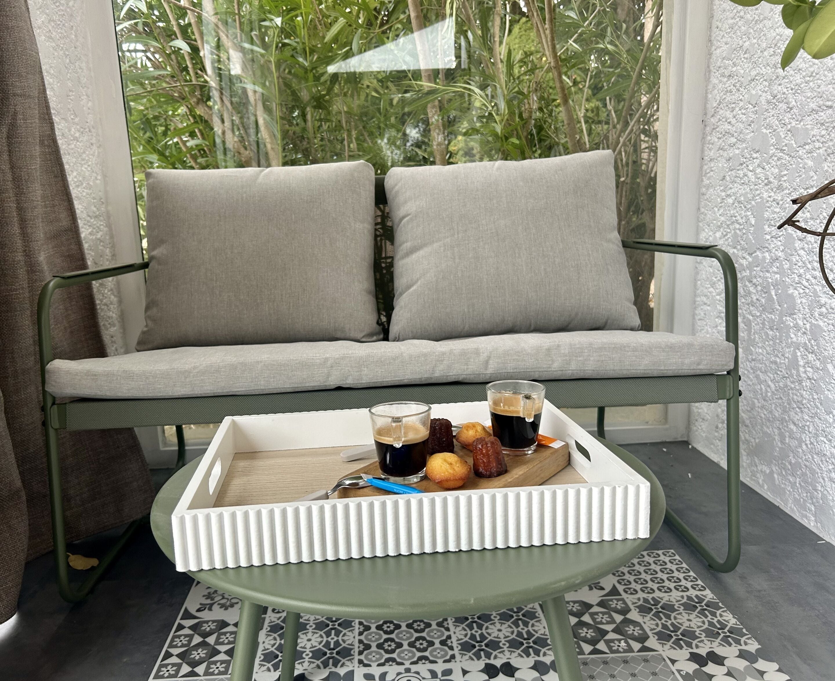 A green metal-framed sofa with two grey cushions, a white tray table with a glass of iced coffee, a pastry, and a spoon.