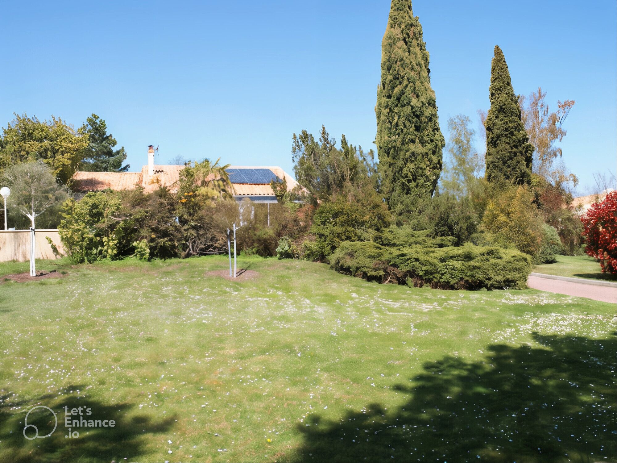 A well-maintained garden with a variety of trees and shrubs, a clear sky, and a building with a solar panel roof in the background.