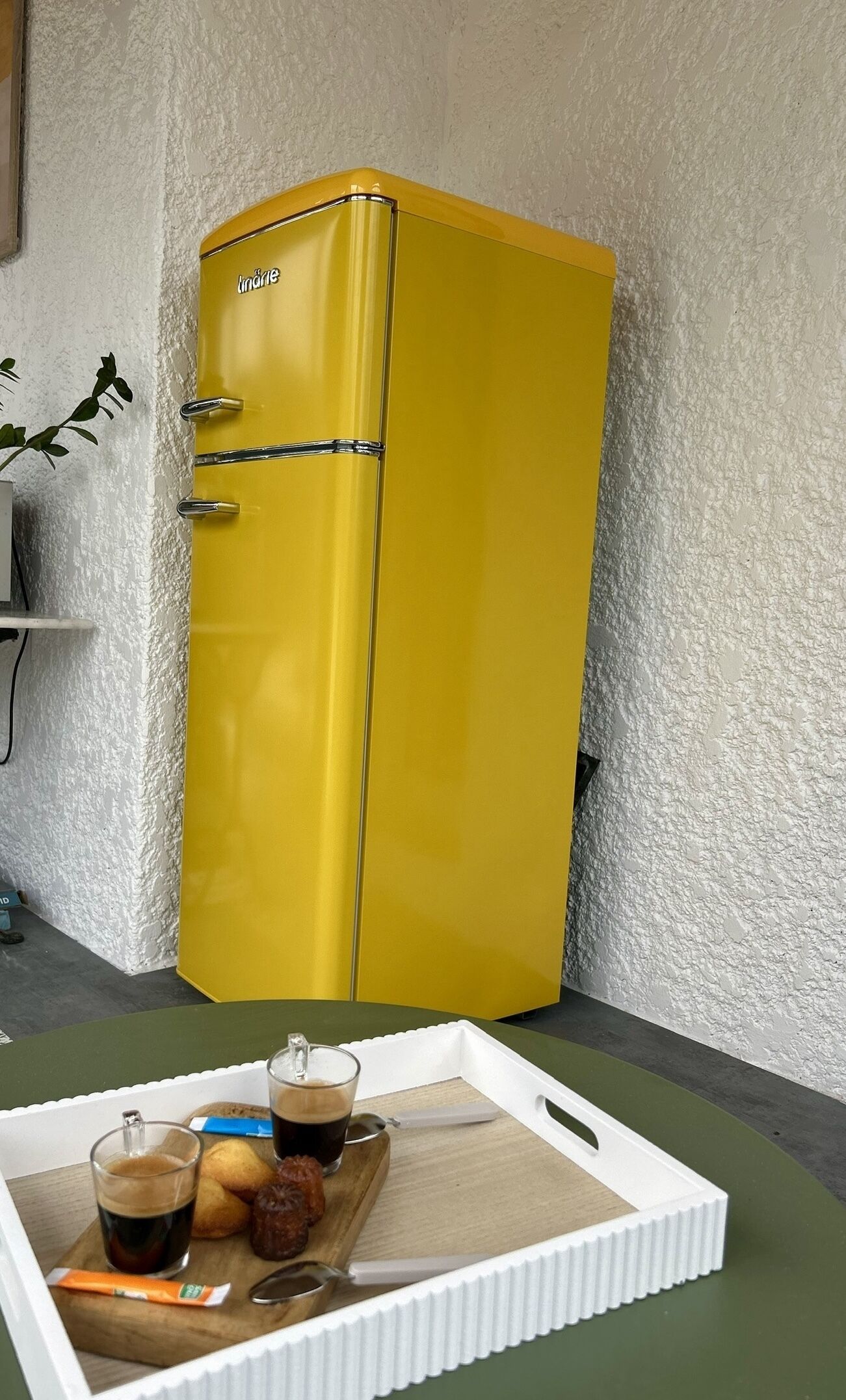 Un vieux réfrigérateur jaune, avec un intérieur blanc, sur une surface en bois. On y voit un plateau avec une tasse de café, une pâtisserie et un sachet de sucre.