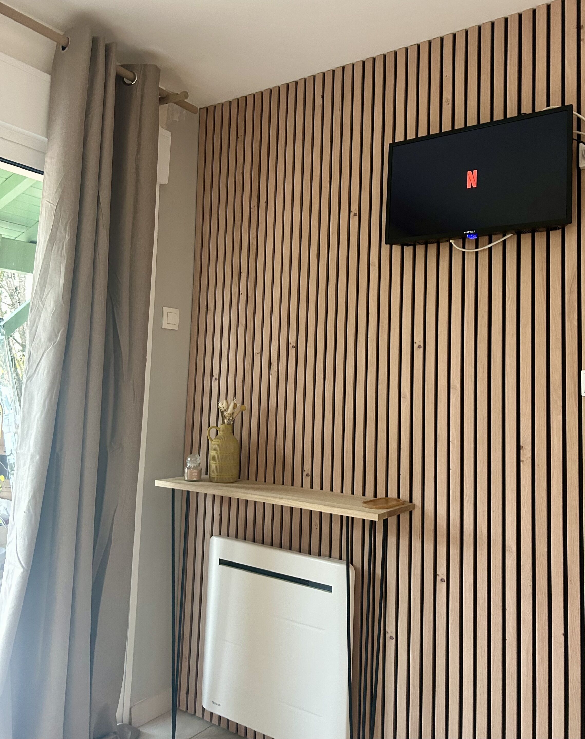Une pièce avec un mur en bois à rayures, un téléviseur fixé au mur, une étagère avec un vase et un radiateur.