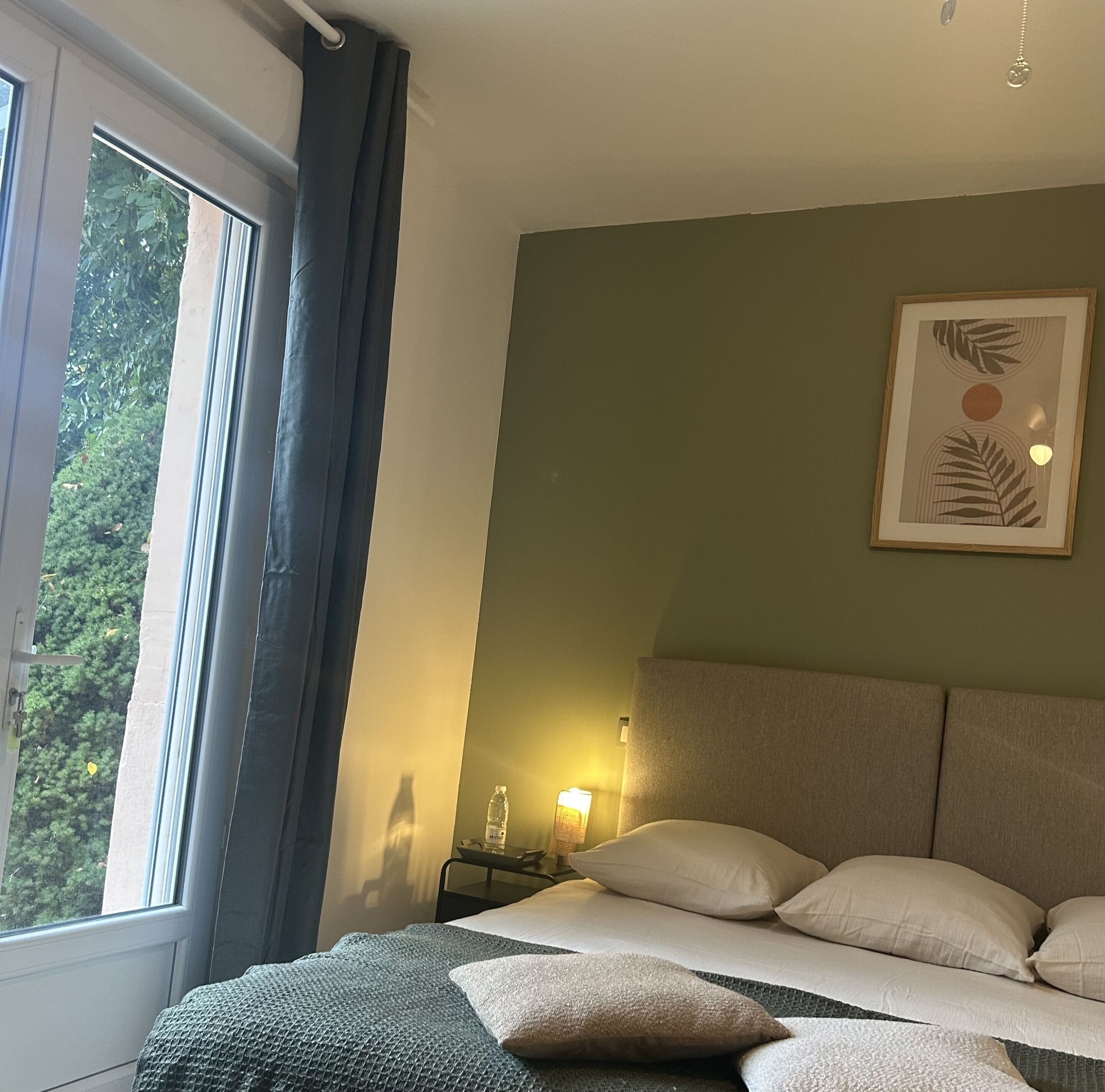 A bedroom with a bed, pillows, a bedside table with a lamp, a framed artwork on the wall, and a window with curtains.