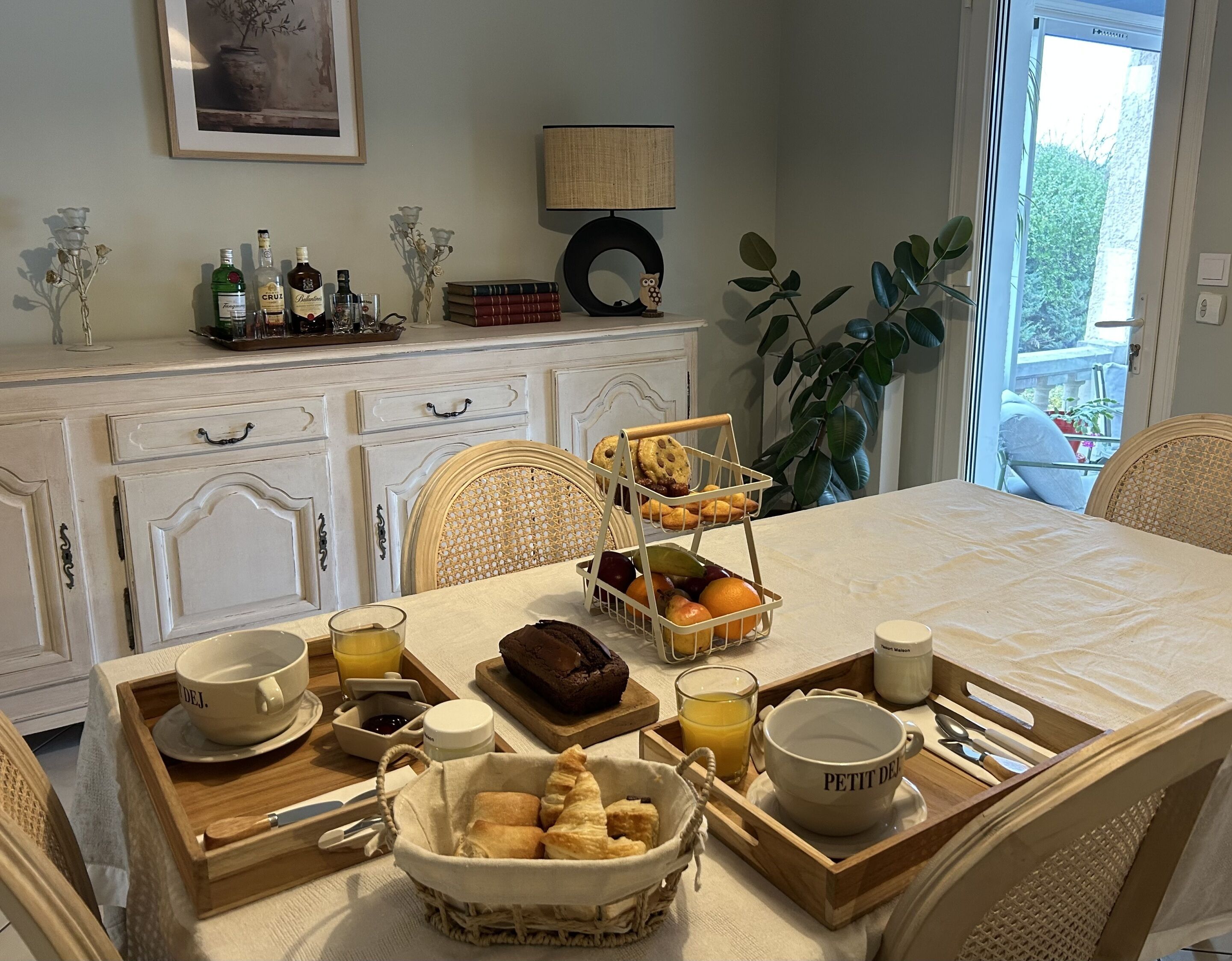 Una mesa de comedor preparada para el desayuno, con pasteles, frutas y bebidas, en una habitación luminosa con vistas al exterior.