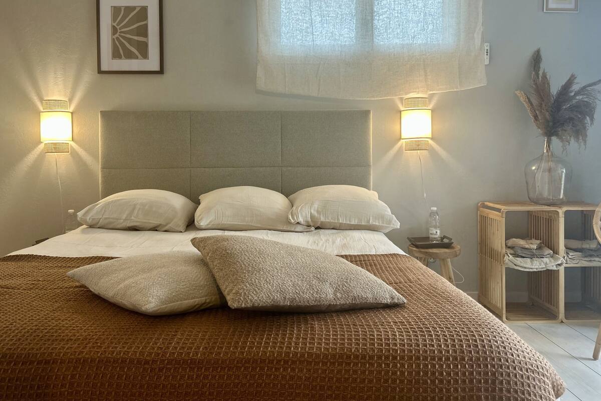 Un lit bien fait, avec une tête de lit, deux lampes fixées au mur, un tableau encadré et une petite table sur laquelle se trouvent un vase et une bouteille.