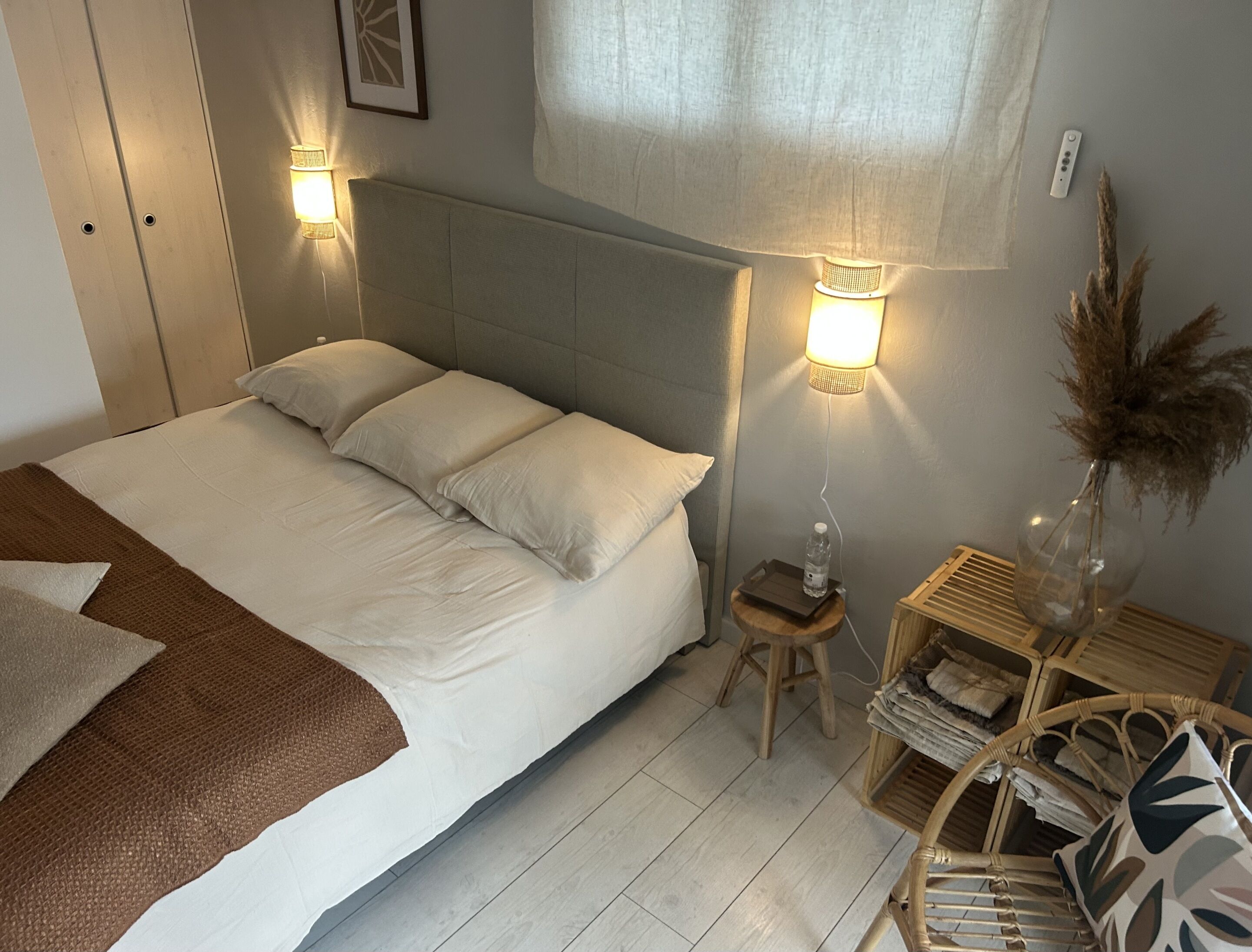 A neatly made bed with white linens and a brown throw blanket, a bedside table with a water bottle, a lamp, and a small stool.