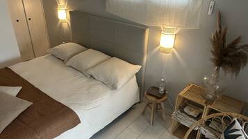 A neatly made bed with white linens and a brown throw blanket, a bedside table with a water bottle, a lamp, and a small stool.