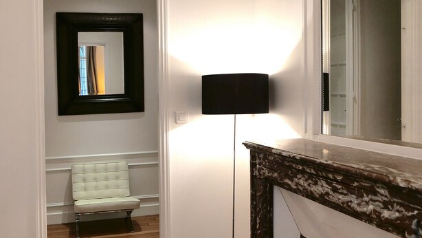 Habitación de hotel con piso de madera, cama blanca, sillón blanco, lámpara de pie negra, chimenea y un espejo grande en la pared.