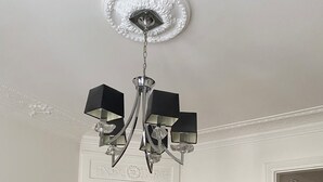 Una habitación con chimenea, un espejo, una araña, un taburete rojo y una pequeña mesa con libros.