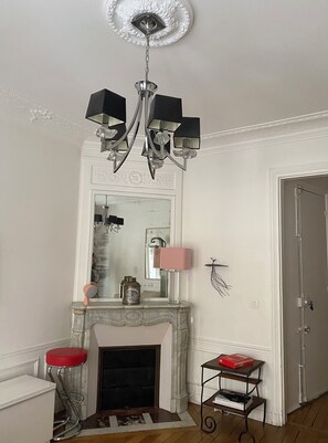 A room with a fireplace, a mirror, a chandelier, a red stool, and a small table with books. - Luxury flat, smart & quiet district (17th) shopping area & pedestrian street (Paris)