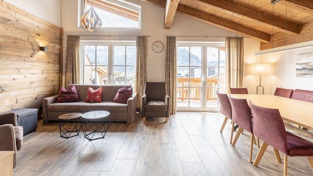 A spacious room with a wooden ceiling, a sofa with cushions, a chair, a dining table with chairs, and large windows offering a mountain view.