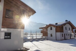 En snedækket gårdsplads med bygninger, der har træfacader og hvide vægge, foran et bjergrigt landskab.