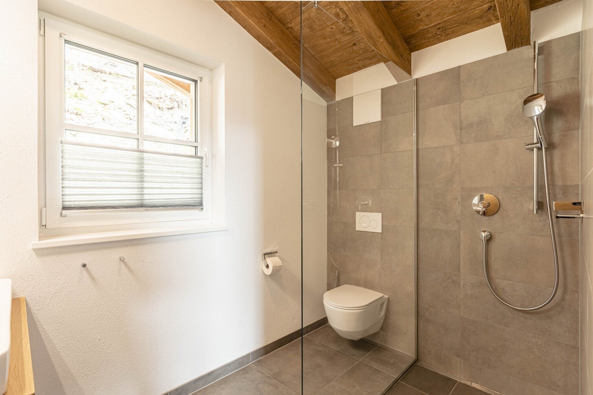 A bathroom with a glass shower enclosure, a toilet, and a window with blinds.