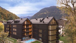 A modern multi-story building with balconies, a swimming pool, and a mountainous backdrop. - Superior 1-Bedroom Apartment (Schruns)