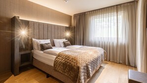 A modern bedroom with a large bed, a headboard with integrated lighting, a bedside table, and a window with curtains. - Superior 1-Bedroom Apartment (Schruns)