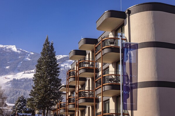 Un hôtel doté de nombreux balcons, avec une vue sur les montagnes et une enseigne publicitaire indiquant « Stations de ski des Alpes ».