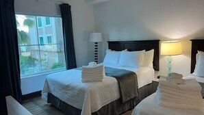 Et hotelværelse med to senge, et stort vindue med gardiner, en sengebordlampe og udsigt til bygninger.