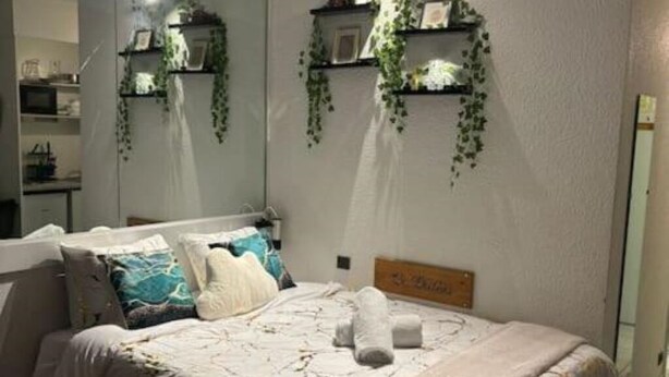 A neatly made bed with a white comforter and pillows, a wooden headboard, and a wall-mounted shelf with plants. - La belle étape and Parking  (Carquefou)