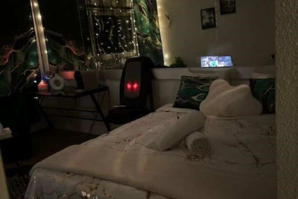 Une chambre à coucher avec un lit, un bureau avec un ordinateur et une chaise. Il y a une fenêtre avec des rideaux et un mur orné d’un tableau.