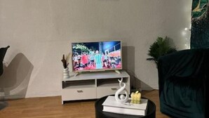 A living room with a television on a white stand, a black side table with a decorative item, and a green armchair. - La belle étape and Parking  (Carquefou)