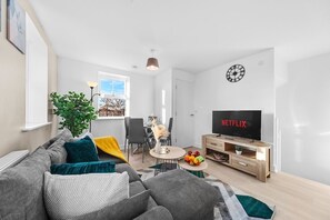 A modern living room with a grey sofa, a coffee table with fruit, a TV, and a dining area with chairs and a table. - Opulent 2BR/2BA House with FREE Parking (Leeds)