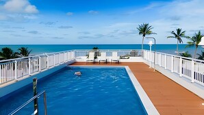 Uma piscina com vista para o oceano, rodeada por um deck com grades brancas e palmeiras.