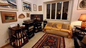 Un salon comprenant un canapé, un téléviseur, une bibliothèque et une fenêtre avec des stores.