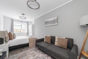 A modern bedroom with a bed, a grey sofa, a patterned rug, and a large window. - Spacious Room In Morden (Morden)