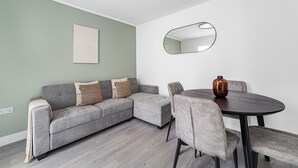 A modern living room with a grey sectional sofa, a round dining table with chairs, a large mirror, and a vase on the table. - Spacious Room In Morden (Morden)