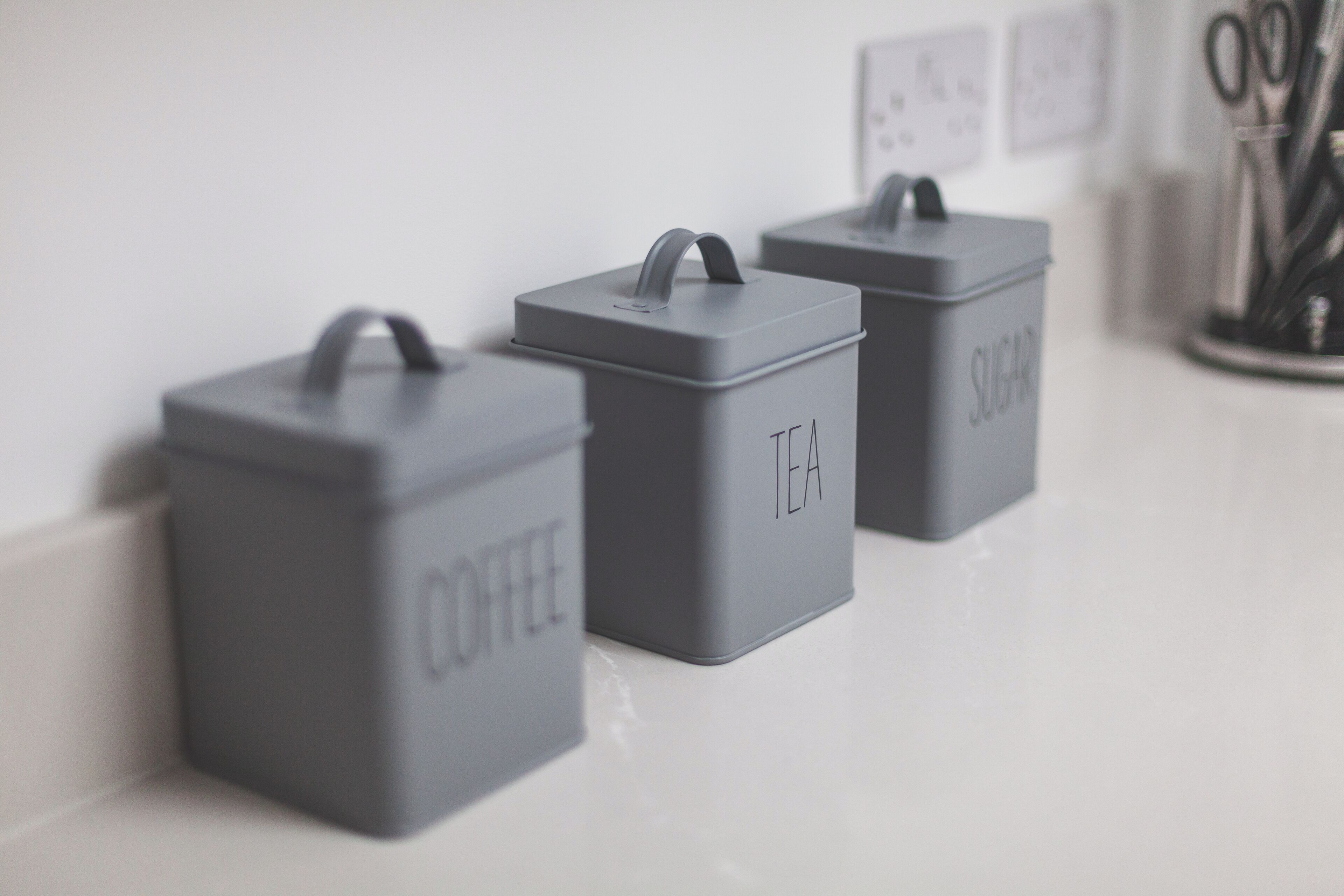 Trois récipients de rangement gris, étiquetés respectivement « CAFÉ », « THÉ » et « SUCRE », sont posés sur un comptoir blanc.