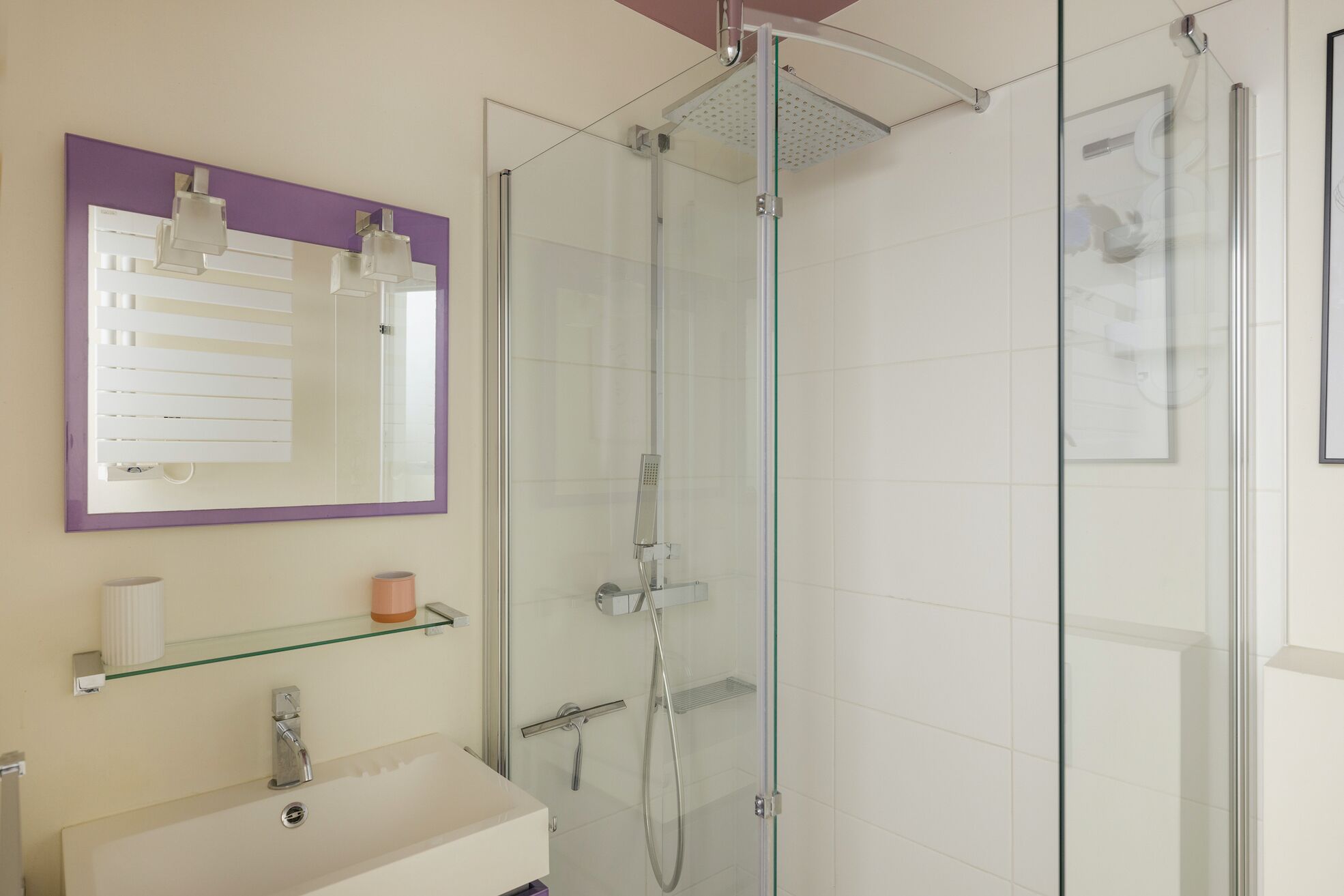 A modern bathroom with a glass-enclosed shower, a white sink, a purple-framed mirror, and a glass shelf with toiletries.
