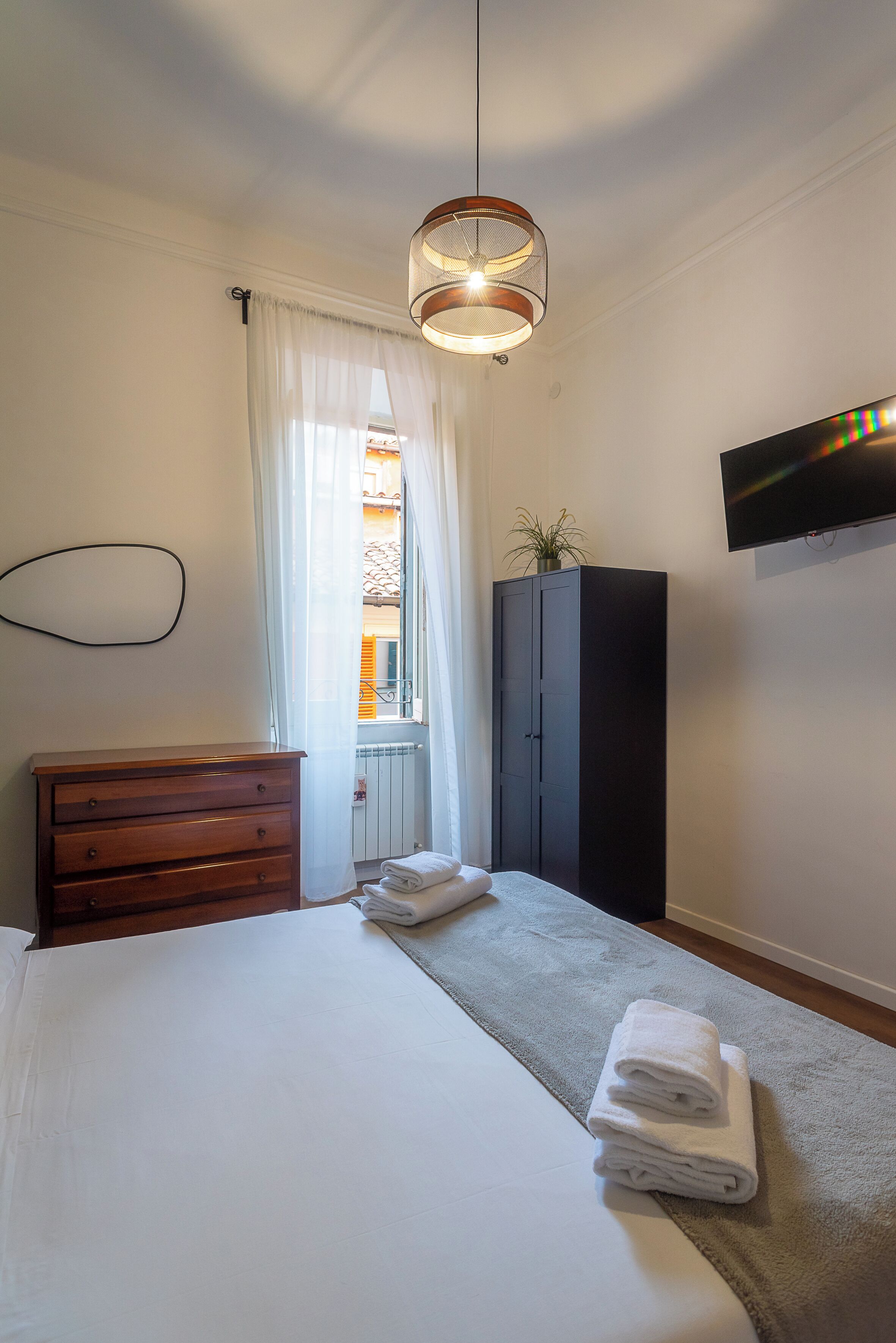A bedroom with a bed, a wooden dresser, a dark wardrobe, a TV mounted on the wall, and a window with sheer curtains.